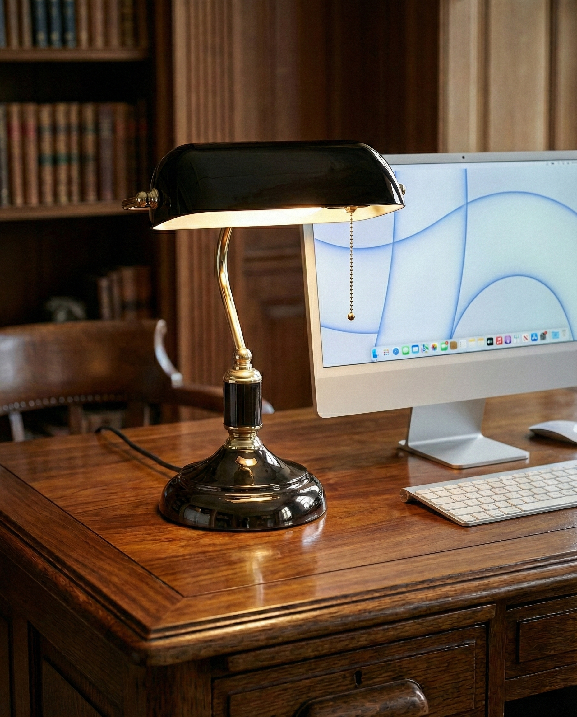 Lampe de bureau style banquier noire avec col de cygne en laiton poli et base noire brillante, allumée, sur un bureau en bois vintage avec ordinateur moderne et bibliothèque en arrière-plan.